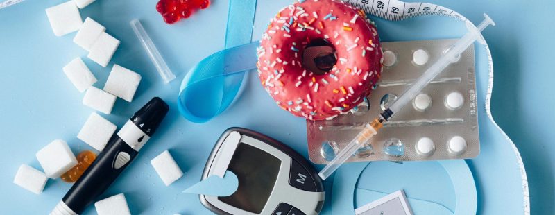 Still life image of diabetes management equipment, sweets, and medication arranged artfully on a blue background.